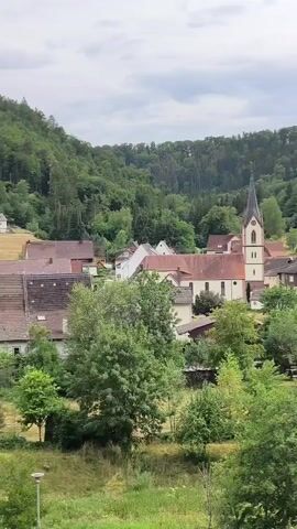A vertical video opens with a view of a small village nestled in a valley. The village features a church with a tall, spire, surrounded by houses with red-tiled roofs. The buildings are situated amongst lush green trees and vegetation. In the background, a densely wooded hill rises up, covered in a variety of green trees. The sky above is overcast with clouds. The video is a static shot with no visible movement.