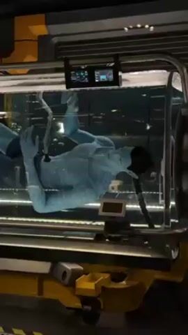 The video shows a clear tank filled with a liquid in what appears to be an industrial setting. Inside the tank, a figure resembling an Avatar from the film "Avatar" floats, connected to various tubes and monitoring devices. The figure is blue with black hair and has a slender physique. The tank is surrounded by yellow and gray machinery, pipes, and monitoring screens displaying data. The atmosphere suggests a laboratory or a specialized facility.