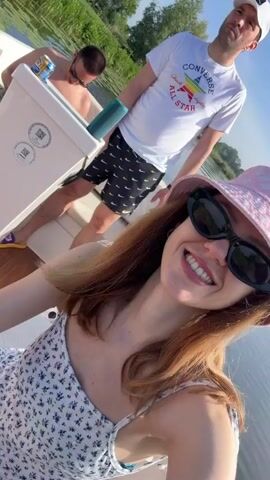 The video shows a group of four people on a small boat, on a river, on a sunny day. A woman with reddish-brown hair, wearing a pink bucket hat and black sunglasses, is holding the camera and smiling. Behind her, two men are sitting and one other woman. One man is wearing a white Converse t-shirt and black shorts, and the other man is shirtless, wearing sunglasses. The other woman is wearing a pink tank top and jean shorts. The boat is moving through the water, with green reeds and lily pads visible on the riverbank. A green portable speaker and beverage are visible.