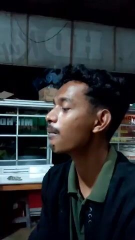 The video shows a side profile of a young man with dark, curly hair, a mustache, and dark skin. He is wearing a dark jacket over a green collared shirt. He exhales a puff of smoke from his mouth. He then brings a cigarette to his lips. The background is dark, but a white, rectangular display case with multiple glass panes is partially visible. Some items are visible inside the case. Above the case are white panels, possibly parts of a ceiling or wall, with some dark lines and writing on them. A table and chair are also partially visible. The lighting is dim.