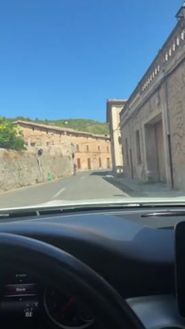 The video shows a view from inside a car, looking forward through the windshield at a narrow, paved street in a village. The street is lined with old, stone buildings with terracotta roofs and wooden shutters. On the left side of the street, there is a stone wall with a tiered structure built into the hillside behind it. On the right side of the street, there are buildings with windows and balconies. The sky is clear and blue, and the sun is shining brightly. In the distance, there is a person riding a bicycle up the street. The dashboard of the car is visible in the foreground, with a steering wheel on the left and a display screen showing "Stevie" and other information.