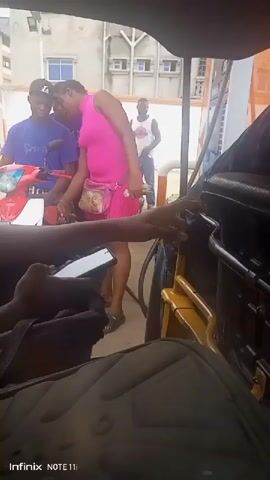 From the vantage point inside a vehicle, possibly a tuk-tuk or auto-rickshaw, the video shows a scene at what appears to be a gas station or fueling area. In the foreground, there's a backpack or bag and an arm holding a black device, possibly a phone or remote. The central focus is on a young woman wearing a pink dress and a fanny pack, standing near a red scooter or moped that a young man in a blue shirt and black cap is attending to. Another man, wearing a white tank top, is standing in the background observing. The surrounding environment suggests an urban setting with buildings visible in the distance. The overall action involves refueling or attending to the scooter while the occupants of the vehicle are observing. The presence of an Infinix NOTE 11 watermark indicates the device used to record the video.