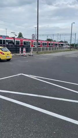 A train with red and white carriages is stopped at a station. The train has many windows and appears to be a commuter train. The station has overhead power lines for the electric train. In the foreground, there is a paved parking lot with white painted lines. A yellow car and a dark gray car are parked in the lot. There are also some temporary orange and white traffic cones visible. The sky is cloudy. The video pans slightly from left to right, showing more of the train and the parking area.