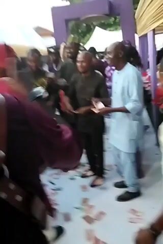 The video shows a group of people gathered at what appears to be an outdoor celebration. Several people are throwing money onto the ground. Many others are milling about and watching the scene. The setting looks like an event venue, with tents or canopies visible in the background. The ground is covered with scattered banknotes. Most people are wearing formal attire. The music is playing in the background.