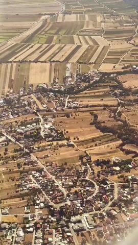 An aerial shot shows a rural landscape with fields and a village. The fields are a patchwork of different colors, indicating various crops or stages of growth, and are divided by lines that suggest roads or property boundaries. A village with houses and streets is nestled within the landscape. The perspective suggests the view is from an aircraft.
