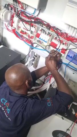 A bald African American man wearing a dark blue work shirt with "Cloud Energy" logo is working on an electrical panel. He is using a tool to connect wires inside the panel. The panel is white and filled with red, blue, and black wires. There are circuit breakers visible. Tools are on the floor to the right.