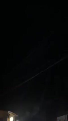 Here's a description of the video:

The video appears to be shot at night on a street lined with buildings. The sky is dark and there are power lines visible. The buildings are lit with warm lights, some with round lamps.  There are a few people walking on the street. Some of the buildings appear to be shops or restaurants. The overall ambiance is quiet and the lighting is low.