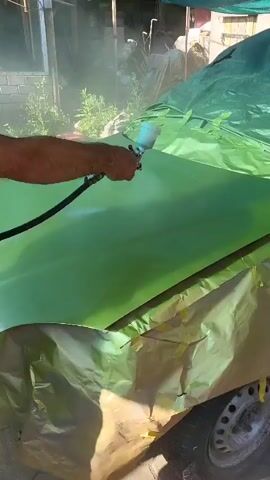 A person's arm and hand are visible holding a white spray gun, painting a green car hood that is partially covered with green and gold plastic sheeting. The spray gun is emitting a fine mist of paint. The background is blurred and shows greenery and structures.