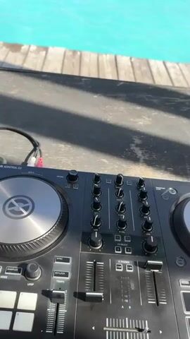 The video shows a DJ controller in the foreground, with a large pool full of people in the background. The pool is surrounded by wooden decking and a canopy structure. The sky is clear and blue. People are swimming, floating on inflatable tubes, and generally enjoying themselves in the water. The DJ equipment suggests that music is being played, likely to enhance the atmosphere of the pool party.