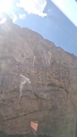 The video shows a person riding in a car alongside a rocky mountain. The car window is quite dirty. The sky is blue with some clouds, and the sun is shining brightly. The camera appears to be handheld and slightly shaky.