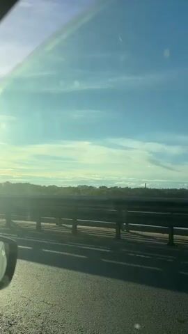 The video is shot from inside a car, looking out the passenger side window. The sun is low in the sky, casting a bright glare and long shadows. The car is on a road that appears to be a bridge, with a metal guardrail visible. Beyond the guardrail, there is a line of trees and some buildings in the distance. The sky is blue with scattered white clouds. The car's side mirror is visible in the lower left corner of the frame.