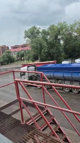The video shows a truck with a blue tarp covering its load. The truck is parked near a facility with trees in the background. A conveyor belt with a brand name "Rakorf" is visible in the foreground, extending over an empty lot where another truck is seen in the distance. The scene appears to be an outdoor industrial area on a cloudy day.