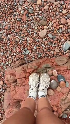 The video shows a rocky beach on a cloudy day.  The ground is covered with small red and grey stones.  In the first shot, a pair of white New Balance sneakers are visible on a larger red rock.  The camera pans up and to the right, revealing more of the beach, some paddleboards, an old barrel, and debris. In the background, there is a body of water with a rock formation and large rocky cliffs.