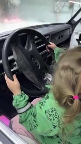 A small blonde girl wearing a green jacket with tigers, pink pants, and pink boots sits inside a white car, holding the steering wheel with both hands. A child in a camouflage jacket can be seen climbing in through the passenger side window. The car's interior is stripped down and partially disassembled, with visible white panels. The steering wheel is black and bears a logo.