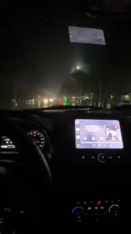 The video shows a view from inside a car driving at night in heavy rain. The windshield wipers are on. Streetlights and other car lights reflect on the wet road. The car's dashboard is visible, including the steering wheel, instrument panel, and a media screen displaying music information. A phone or similar device is mounted on the windshield.