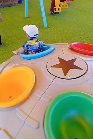 A young child wearing a white baseball cap with the number 23 and a colorful Hawaiian shirt is sitting in a blue seat on a circular, gray playground structure. The structure has various colored compartments, including yellow, red, and green. The child is looking to their left and has their legs extended. The playground is on a green, artificial turf surface.