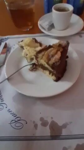 A slice of chocolate cake with nuts is on a white plate in the foreground. A fork is stuck in the cake. To the left of the plate is a knife. Behind the plate is a placemat with blue lettering. To the right of the plate, on a saucer, is a small white cup of espresso. Next to the espresso cup are two cans of soda. To the left of the espresso cup is a glass of iced tea. In the background, on an orange table, is a painting of a woman with a colorful headwrap.