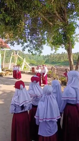 A group of young schoolgirls in white headscarves and burgundy skirts stand in a circle outside, holding hands.  In the background, a group of schoolgirls, also in uniform, stand on a wooden platform.  Behind them is a river and a lush, green landscape.  A large tree provides shade over the circle of girls.  It is a sunny day.