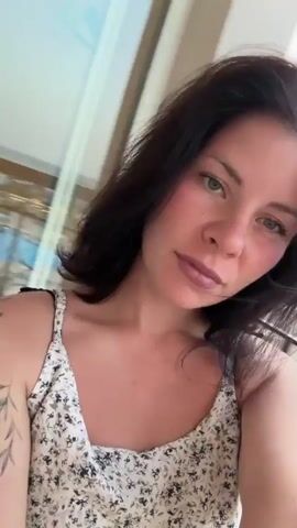 A fair-skinned woman with dark hair and light eyes is the subject of this selfie-style video. She's wearing a white tank top with a small floral pattern, and a tattoo is visible on her left arm. Her face is flushed, possibly from heat or exertion, and she's wearing mauve lipstick. The background includes a neutral-colored wall with large glass doors or windows. The audio has music playing. The camera is handheld and moves slightly, capturing her from various angles. She briefly runs her hand through her hair and purses her lips.