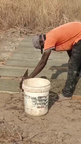 A slender, dark-skinned man in an orange t-shirt, dark-colored pants, and a baseball cap is working on a construction project outdoors. He's bent over, using a trowel to apply a gray mortar-like substance to what appears to be a low wall or foundation made of gray blocks.  A white plastic bucket sits near him on the dirt ground. He wears a dark glove on his working hand and stands barefoot on the bare ground.  Dry grass and vegetation are visible in the background.