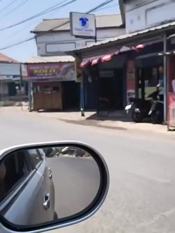The video shows a street scene with several small businesses. In the foreground, the perspective appears to be from a moving vehicle, possibly a motorcycle, as a side mirror and handlebars are visible on the right. A street sign for "Seles Laundry" is visible on the left. Further down the street, there are storefronts. One storefront displays a sign that says "AVOCADO DURAN." Another storefront has a sign that reads "KEBAB DAN BURGER QIU QIU." A traffic cone is placed on the side of the road in front of one of the businesses. The businesses appear to be open, with some displaying menus or food items. There are motorcycles parked in front of some of the establishments. The environment is an outdoor urban setting on a sunny day.