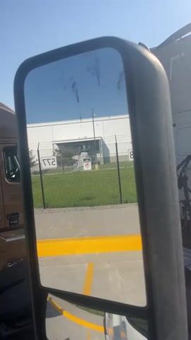 This media file shows a reflection in the side mirror of a truck. The reflection displays a large, white industrial building in the background, with a chain-link fence and green grass in front of it. A sign with a circular red symbol and a diagonal line is visible on the fence. In the foreground of the reflection, there's a yellow line painted on the ground, indicating a parking space or a boundary. The sky is clear and blue, suggesting a sunny day. Part of a brown truck is visible on the left side of the frame, and another white truck is partially visible on the right. The overall scene appears to be an industrial or commercial area, possibly a loading dock or a parking lot, given the presence of trucks and the industrial building. The time of day is likely midday due to the bright sunlight and clear sky. There is no visible text to decipher, apart from what might be painted on the trucks or signs, which are not clearly legible in the reflection. The overall mood is neutral, depicting a typical day in an industrial setting.