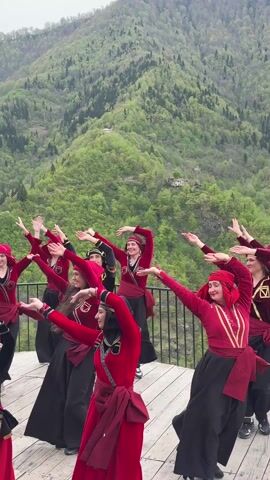 A group of women and one girl are dancing on a wooden platform in front of a mountain covered in trees. The women are all wearing matching red velvet tops with gold embroidery, black skirts, and red hats. They also have red belts tied around their waists. The girl is wearing a similar outfit, but her hat is pink. The dancers are arranged in a semi-circle. They are all smiling and looking at the camera as they perform the dance. The backdrop is a lush green mountain with a few houses visible. The sky is overcast. The camera remains steady throughout the shot.