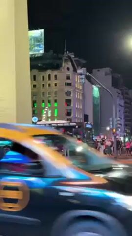 The video depicts a bustling city scene at night, with a prominent white obelisk towering in the background. Cars and a bus traverse a crosswalk in the foreground, while pedestrians stroll along the sidewalks. Buildings with illuminated windows and advertisements flank the obelisk, contributing to the vibrant urban atmosphere. The sky is dark, with a visible moon adding to the nighttime ambiance.