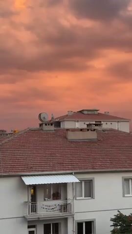 A video shows a view of buildings with red-tiled roofs against a vibrant orange and pink sky filled with clouds. The foreground features the top of a white building with a balcony and windows. Behind it are other buildings with similar roofs, and further in the distance, larger apartment buildings are visible. In the far distance, mountains can be seen. The overall impression is a sunset over a residential area.