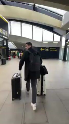 A man is walking through an airport terminal pulling two suitcases. He is wearing a black coat, grey pants, white sneakers, and a black backpack. The suitcases are black and white. He is walking towards a sign that says "Airport terminals" with an arrow pointing to the left. Other people with luggage are visible in the background. The terminal has large windows, a high ceiling, and tiled floors.
