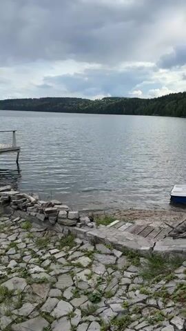 A drone captures a view of a calm lake on a cloudy day. The shore in the foreground is paved with rough stones and some patches of grass. A wooden dock extends into the water on the left. On the right, a small white boat is docked near a wooden ramp. The far shore is lined with a dense forest.
