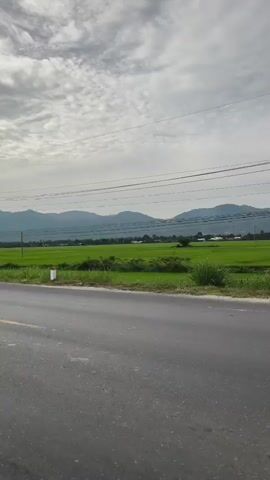 The video is a landscape shot of a road in front of green fields. The sky is overcast. In the distance, there are mountains. There are power lines running across the sky. The road is asphalt and has a yellow line down the middle. There are white and red bollards along the side of the road. A motorcycle with two people on it rides by.