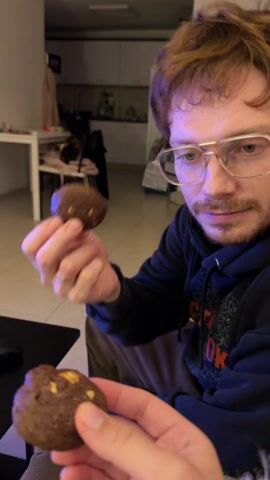 A man holds up two chocolate cookies in front of the camera. He is wearing glasses and a dark blue hoodie with a design on the front. He looks at the camera while holding the cookies. The person holding the camera reaches out and takes one of the cookies. The background shows a partially visible room with white walls and a few pieces of furniture.