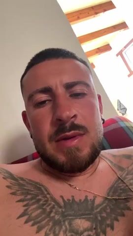 The video features a man indoors, chest bare, with a beard and short hair. He has a large tattoo on his chest, and a gold chain around his neck. The background includes a wall, part of a ceiling with exposed beams, and a plaid pattern which may be a blanket or part of a sofa. The man closes his eyes briefly.