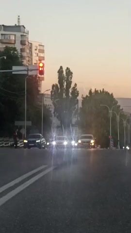 The video captures a street scene in Pyatigorsk, Russia, during what appears to be either dawn or dusk due to the soft, diffused light and the warm hues in the sky. Several cars are on the road, with headlights on, suggesting low light conditions. One traffic light is visibly red.

Pedestrians are crossing the street at a marked crosswalk. Some individuals are walking in groups, while others are alone. A white car is in the process of turning right. The background features apartment buildings and trees, typical of an urban environment. The overall mood is calm, with no apparent distress or urgency.

The weather seems clear and calm, with no signs of precipitation. The presence of streetlights and traffic signals indicates an active urban area. The video does not contain any visible text.