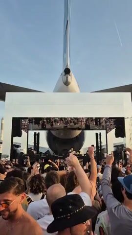 The video captures a lively outdoor music event with a large crowd in front of a stage, strikingly framed by the tail of an airplane. The crowd is dense, with many people raising their hands in the air, seemingly enjoying the music. The stage is visible through a rectangular structure and has speakers. The backdrop behind the stage is dominated by the large tail section of a white and blue aircraft. The sky above is clear. The video is shot from within the crowd, providing a first-person perspective of the event.