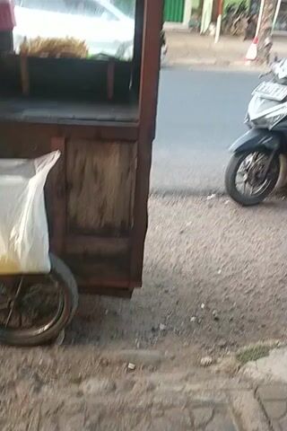 A street scene is captured in portrait orientation. In the foreground, a wooden food cart with glass panels and metal accents is positioned on the right side of the frame. A motorcycle is parked to the left of the food cart, with its front wheel on the paved road and its rear wheel near the cart. A person wearing a light-colored shirt and a white helmet is seated on the motorcycle, facing the food cart. The food cart has a wooden door on the right side, and its interior appears to contain food items. Behind the food cart, a building with a green shuttered window is visible, and several motorcycles are parked in front of it. Flags are also present, including what appear to be Indonesian flags. The ground is paved with asphalt and has some loose gravel and dirt. The scene suggests a vendor operating a food cart on a street.