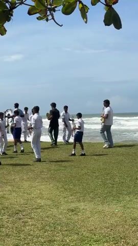 A group of young people dressed in white uniforms and some in dark green uniforms are standing on a grassy area near the ocean. Some are holding musical instruments, including trumpets and what appears to be a snare drum. They appear to be preparing for or in the middle of a musical performance. The ocean is visible in the background with waves rolling in. Trees with green leaves are in the foreground, partially obscuring the view. The sky is blue with some clouds.