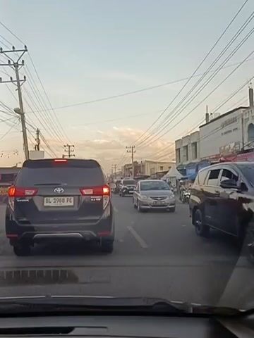 The video captures a traffic scene on a street. Several cars are visible, including a black SUV in the foreground with the license plate "BL 1988 PE." A red car is maneuvering in front of the SUV, and other vehicles, including a motorcycle with two riders, are present further down the road. Buildings line the right side of the street, some with awnings and signage. Power lines stretch across the sky, and utility poles are visible on the left. The sky is partly cloudy, suggesting it might be late afternoon or early evening. The overall impression is that of moderate traffic congestion.