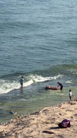 The video shows a beach scene with a body of water. There are several people visible. In the water, near the shore, a person is standing. Further out, two people are interacting with a large, cylindrical object. Another person is visible in the water closer to the shore, and a fourth person is standing on the sand. The environment appears to be a sunny day. The sand in the foreground shows some debris.