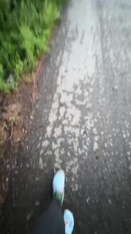 The video begins with a close-up of a person's hand with white nail polish reaching towards a blue Croc shoe with a red and green accent. The person is wearing a black hoodie and gray leggings. The ground appears to be wet pavement with some debris. The camera then pans upwards and blurs, showing a bright sky and trees. The video continues with the camera spinning and focusing on the person's face, who is looking at the camera with a surprised expression and puckered lips. The person is wearing a black hoodie with a white drawstring and has long, light brown hair. The background consists of trees and a blue sky. The scene appears to be outdoors in daylight.