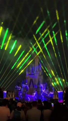 The video shows a nighttime scene in front of a castle. A crowd of people are gathered in the foreground, facing the castle. The castle is illuminated with colorful lights and projections. Green and yellow laser beams crisscross the sky above the castle. Some of the projections on the castle appear to be animated images. The sky is dark, indicating it is nighttime. There are no discernible actions or interactions taking place within the crowd, other than observing the show. The overall environment is that of an outdoor nighttime event or performance.