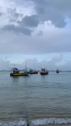 This video shows several fishing boats on a body of water under a cloudy sky. The water appears to be the ocean, with small waves breaking on a sandy shore in the foreground. The boats are of various sizes and colors. The closest boat to the left is yellow, blue, and green, with a red roof. It has fishing gear, including poles and nets, attached. To the right of this boat, there are two smaller red boats. Further in the distance, on the far right, is another red boat. The sky is filled with dark, moody clouds, suggesting overcast weather or an approaching storm. The overall scene is tranquil and evokes a sense of the open sea.