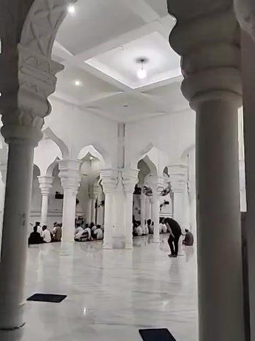 The interior of a white mosque is shown in the video. The architecture features rows of white columns supporting arched openings. People dressed in religious attire are sitting or walking around, likely engaging in prayer or religious activities. The floor is a polished, reflective surface, and ceiling lights illuminate the room. Several ceiling fans are visible. A person stands alone in the room.