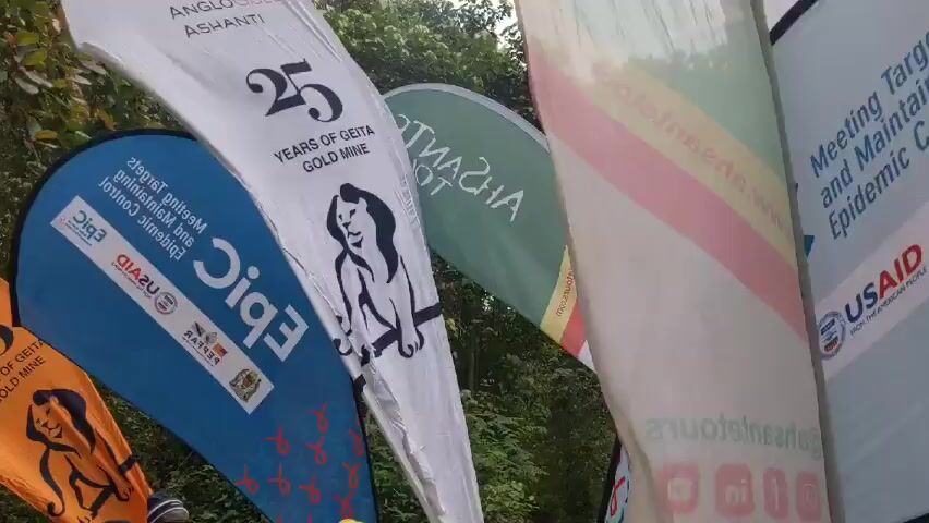 A group of people are gathered outside, many wearing yellow safety vests, and some wearing helmets. They are holding up several flags and banners with various logos and text. The banners include the words "TANZANIA COFFEE BOARD" and "GGM Kili Challenge Crew".  Some of the people are cheering and singing. The setting appears to be a wooded area.