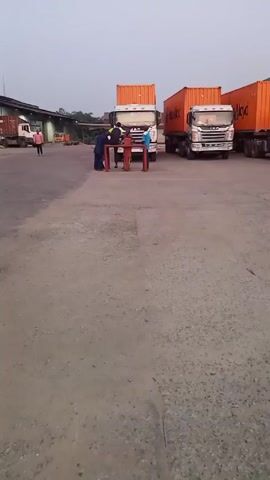 The scene is an open, paved lot possibly associated with shipping or transportation, based on the presence of several large semi-trailer trucks. Some trucks are carrying shipping containers. Along one side of the lot is a long, low building, potentially a warehouse or depot. A few individuals are visible. One person is walking across the lot, and a small group of people is gathered near a table set up in front of one of the trucks. The sky is overcast and the light suggests it's either early morning or late evening. The ground is cracked and shows signs of wear.