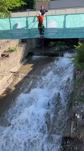 The video captures an outdoor scene featuring a bridge spanning over a fast-flowing river or stream. The river cascades down several man-made steps, creating whitewater rapids. The banks of the river are reinforced with stone gabion walls and concrete structures, with lush greenery visible on the upper banks.

Two men are visible in the frame. One man, positioned centrally, is wearing an orange long-sleeved shirt, dark pants, a white helmet, and a climbing harness. He stands on a narrow platform or rope system, with ropes extending downwards, indicating he is preparing for a jump (likely a rope jump or bungee). His arms are outstretched, and he appears focused on the activity. Another man, dressed in a dark shirt, stands on the bridge railing directly behind the jumper, observing the preparations.

The bridge itself has a turquoise or light blue barrier with white text in Cyrillic. Two distinct phrases are visible: "КРАСНОДАРСКИЙ КРАЙ" (Krasnodar Krai) and "РУССКИЙ СЕВЕР" (Russian North). The setting appears to be during daytime, with clear weather, indicated by strong shadows cast on the river banks and water. The overall impression is one of an adventurous activity taking place in a scenic, developed natural environment.