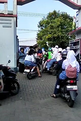 The video shows a busy scene with many people, mostly young women wearing school uniforms and headscarves, on and around motorcycles. They are gathered near an archway or gate, possibly entering or leaving a school or other institution. The ground is paved with cobblestones. Several motorcycles are parked, and others are in motion. Some individuals are wearing helmets, while others are not. The overall atmosphere appears to be one of transition, with many people seemingly on their way somewhere.