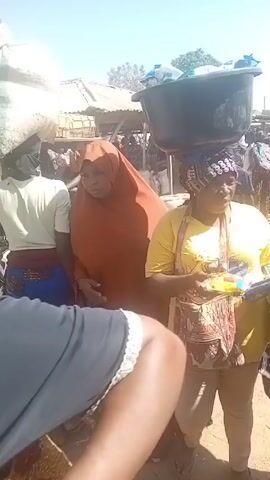 The video shows a bustling outdoor market scene. Several people are walking around, some carrying large items. One woman has a large black basin balanced on her head, while another wears a brown hijab and carries a snack. Men are pulling wheelbarrows laden with goods. The market appears to be sandy and sunny.