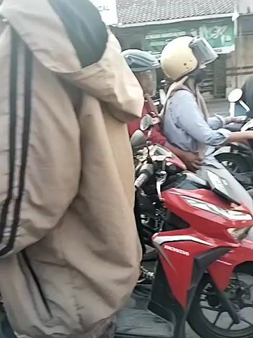 The video shows a street scene with several motorcycles and a car. There are multiple people on the motorcycles, most of whom are wearing helmets. One person in the foreground is wearing a black hoodie and jeans, sitting on a red motorcycle. Another person behind them is wearing a pink shirt and jeans, also on a motorcycle. Further back, a person in a light blue shirt and jeans is riding a motorcycle, with another person behind them wearing a light-colored top and dark pants. A silver car is stopped in the middle of the road, with people on motorcycles around it. The environment appears to be an urban or semi-urban street, possibly in a warm climate, with buildings and trees visible in the background. The overall impression is of a busy street with traffic.